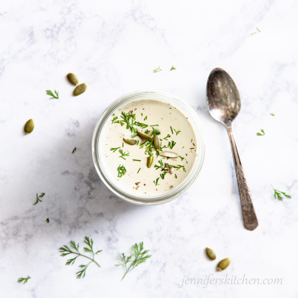 Creamy Pumpkin Seed Dressing in a jar garnished with herbs and pumpkin seeds with a spoon on the side.