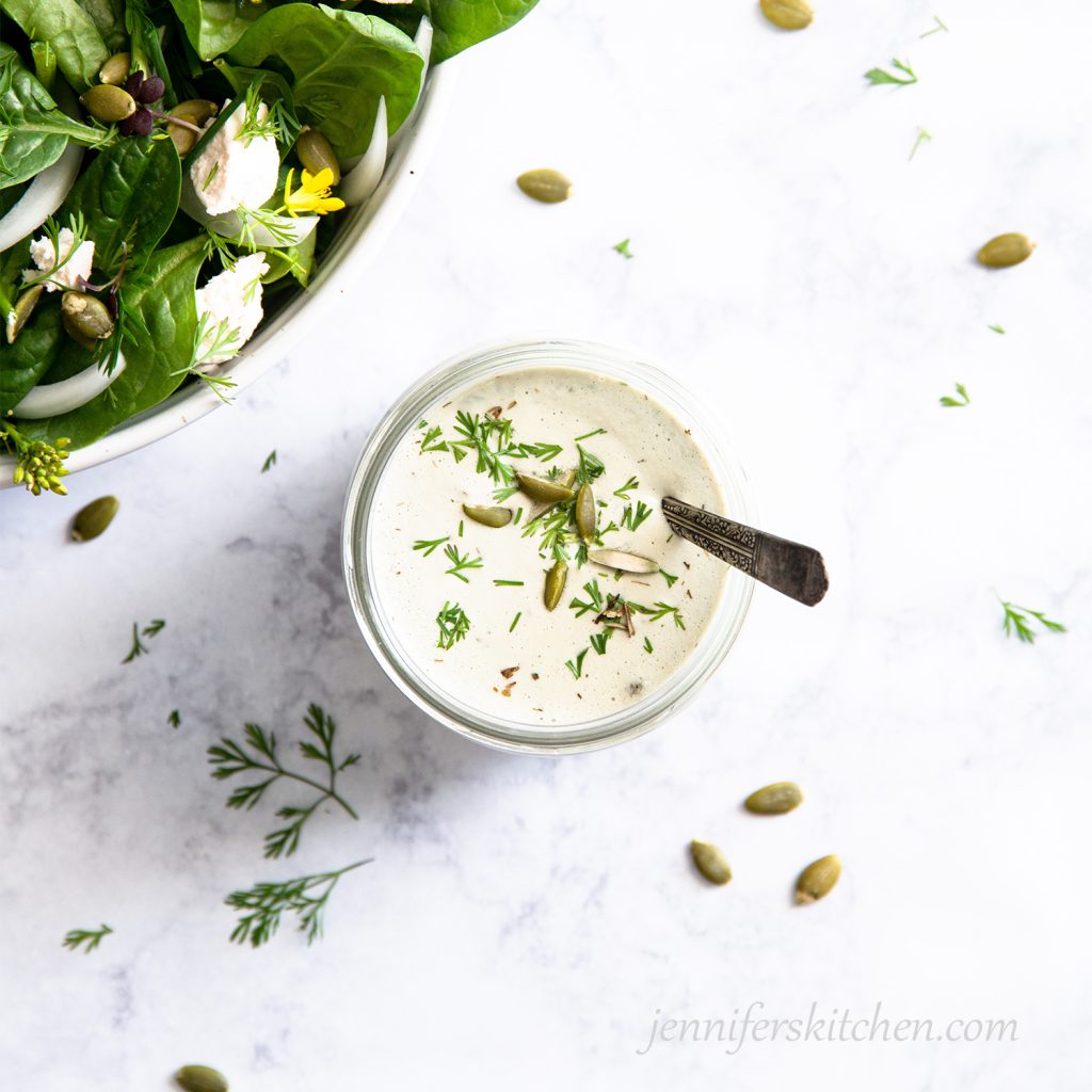 Creamy Pumpkin Seed Dressing in a jar with a spoon garnished with herbs and pumpkin seeds with a salad on the side.