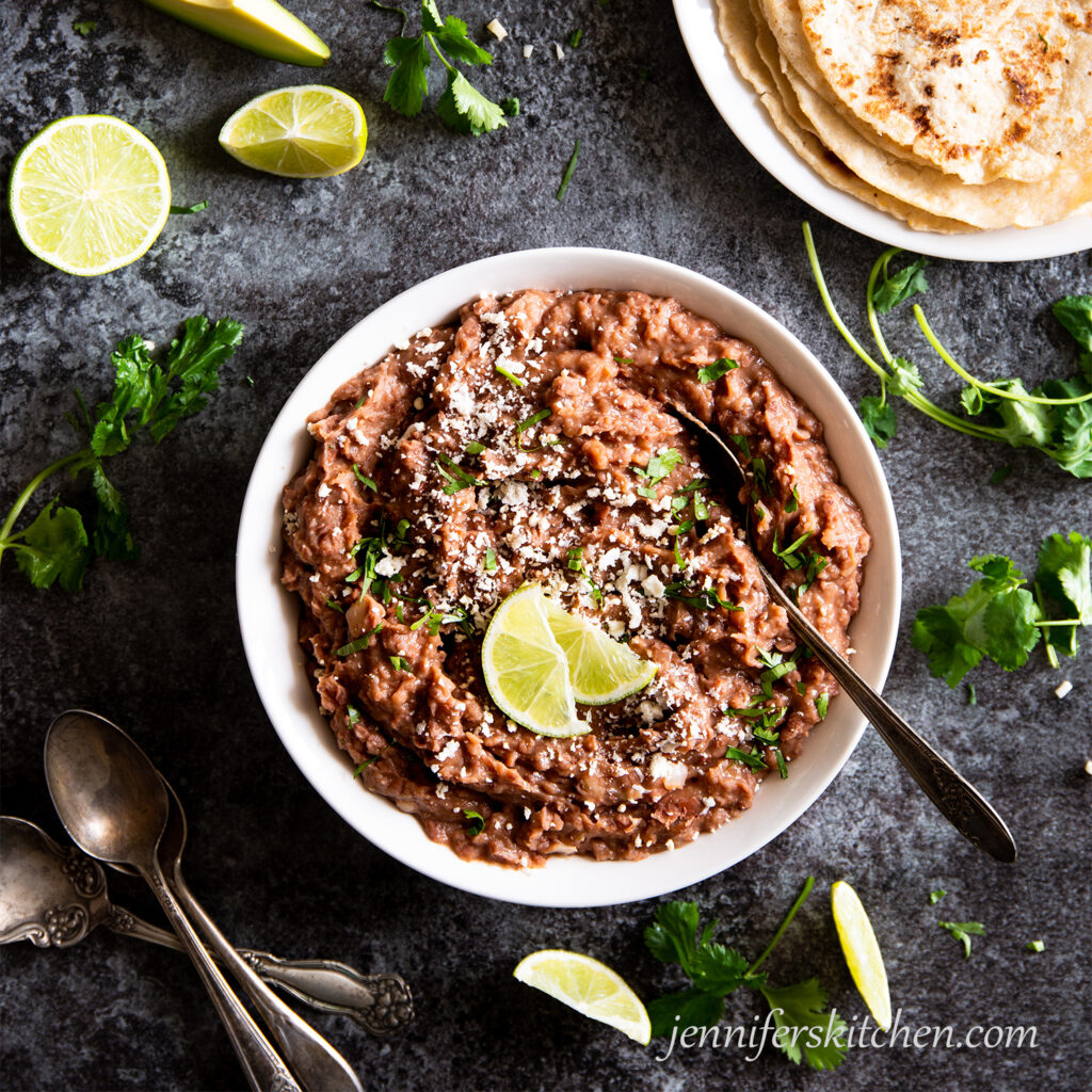 A bowl of refried beans topped with minced garlic, cilantro, and lime wedges.
