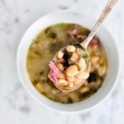 Top view of a spoonful of smoky white bean soup with ham and the white bowl on a marble background