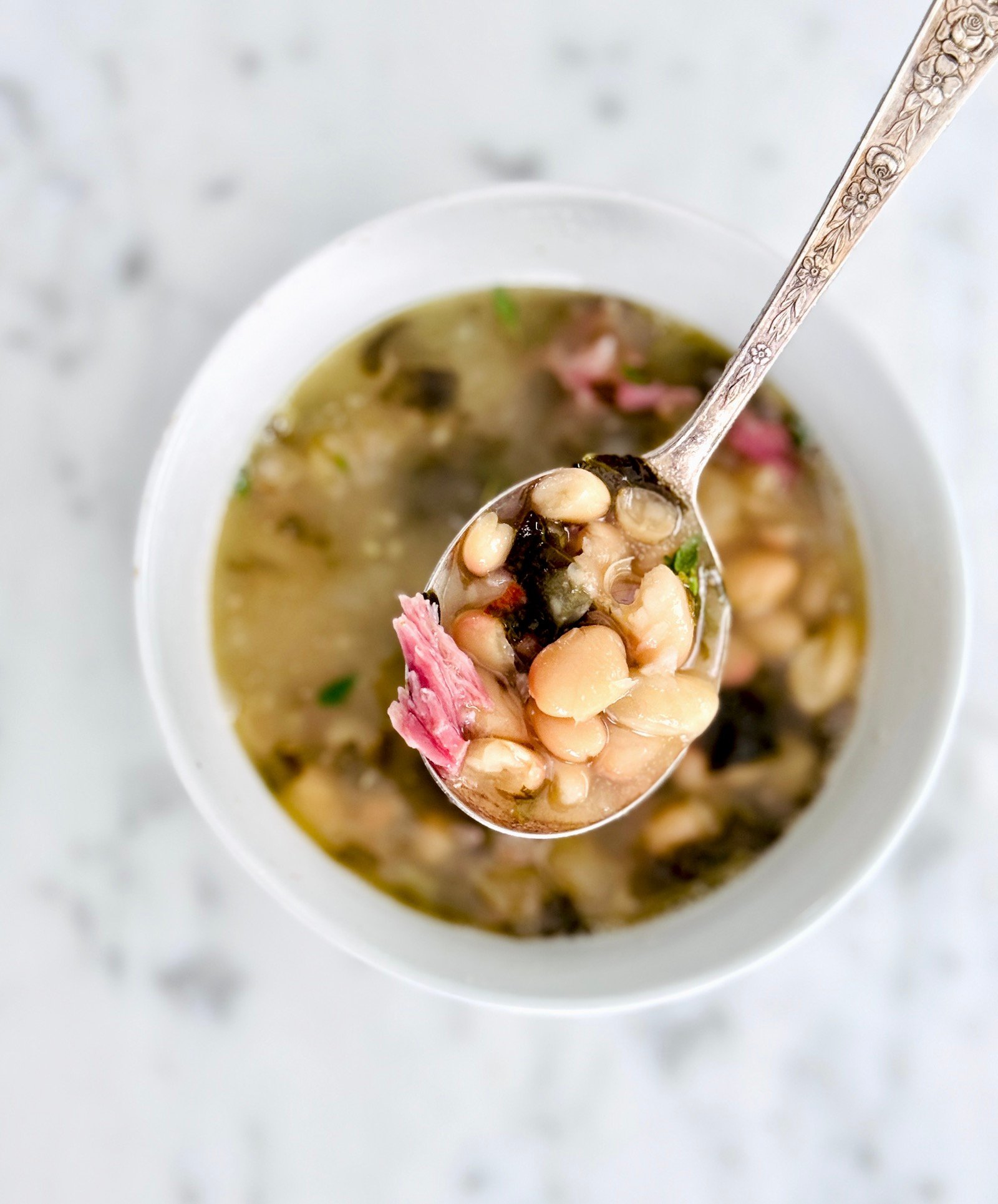 Top view of a spoonful of smoky white bean soup with ham and the white bowl on a marble background